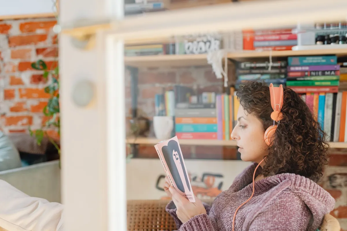 Vrouw met koptelefoon leest rustig een boek bij een boekenkast