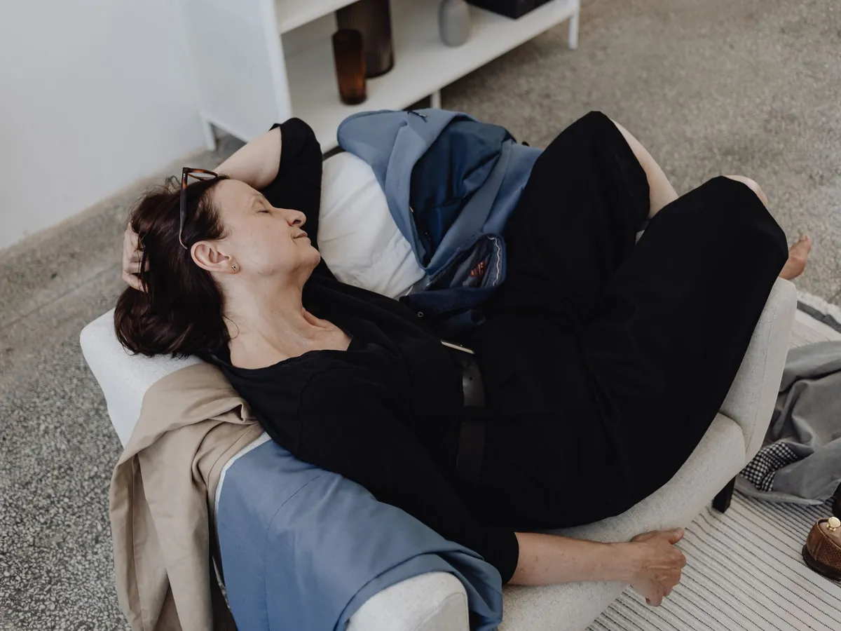 Vermoeide vrouw ligt languit op een bank in een lichte woonkamer met kleding om zich heen