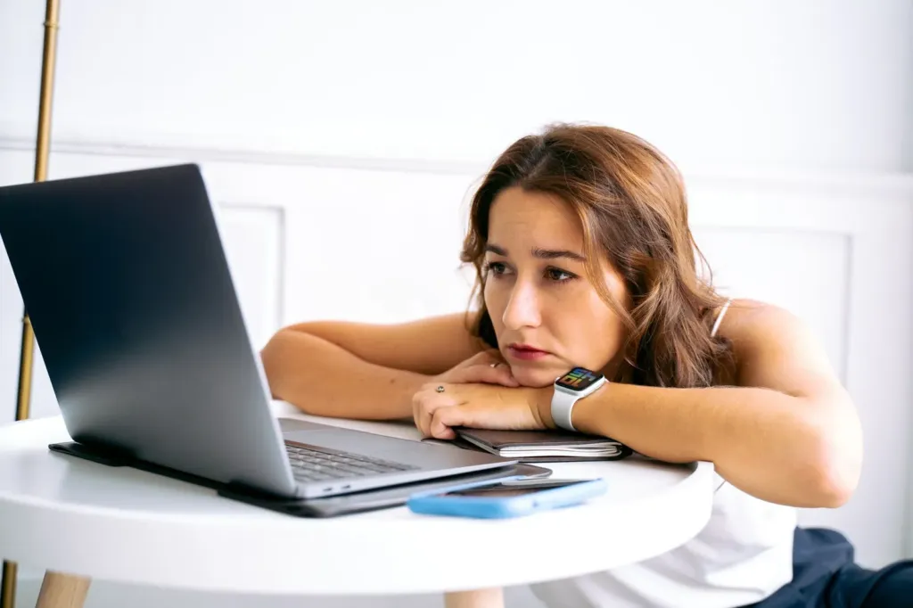 Vermoeide vrouw leunt met haar armen op een tafel en kijkt naar een laptop