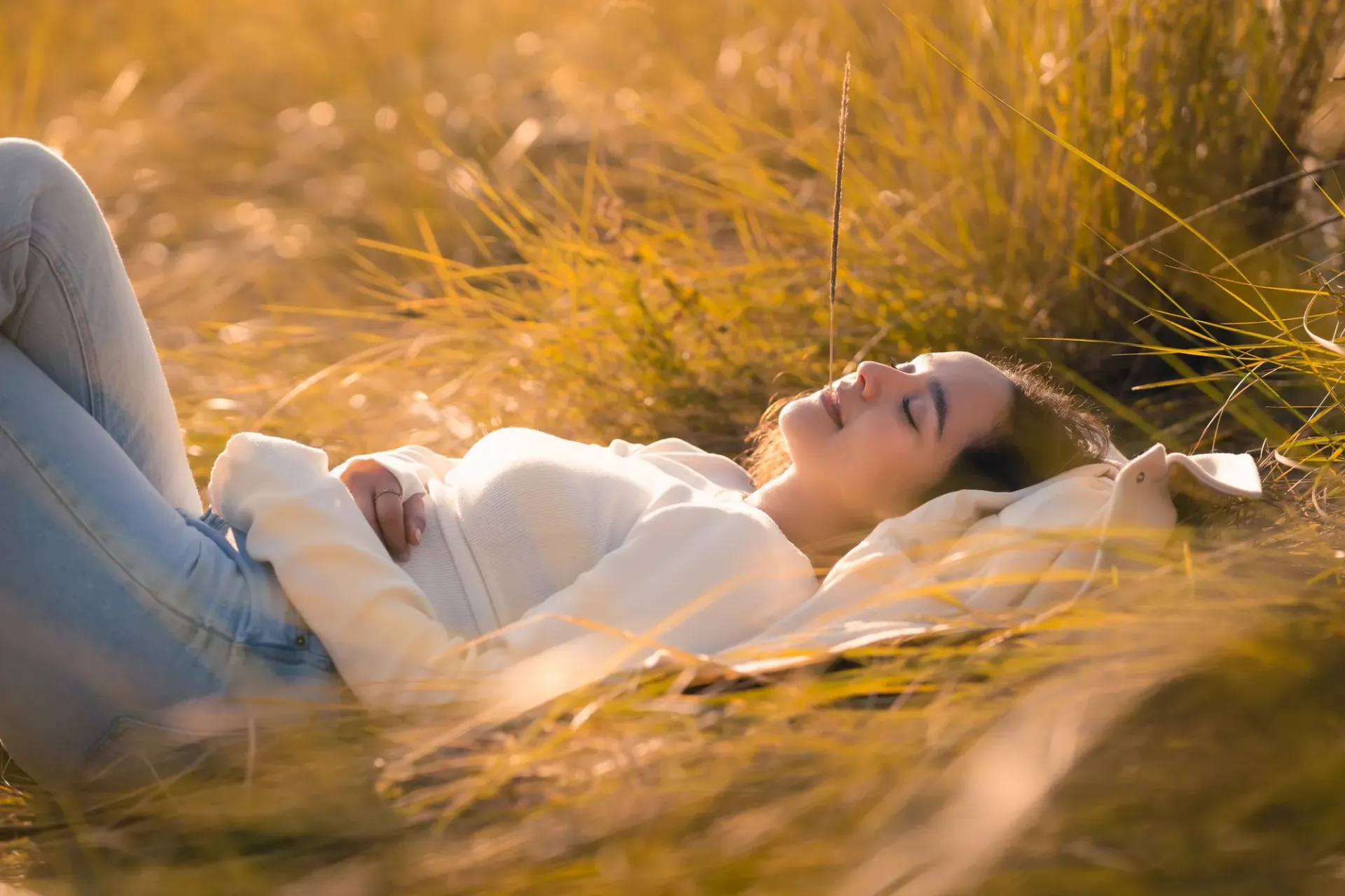 Vrouw ligt ontspannen met gesloten ogen in hoog gras dat oplicht in het zonlicht
