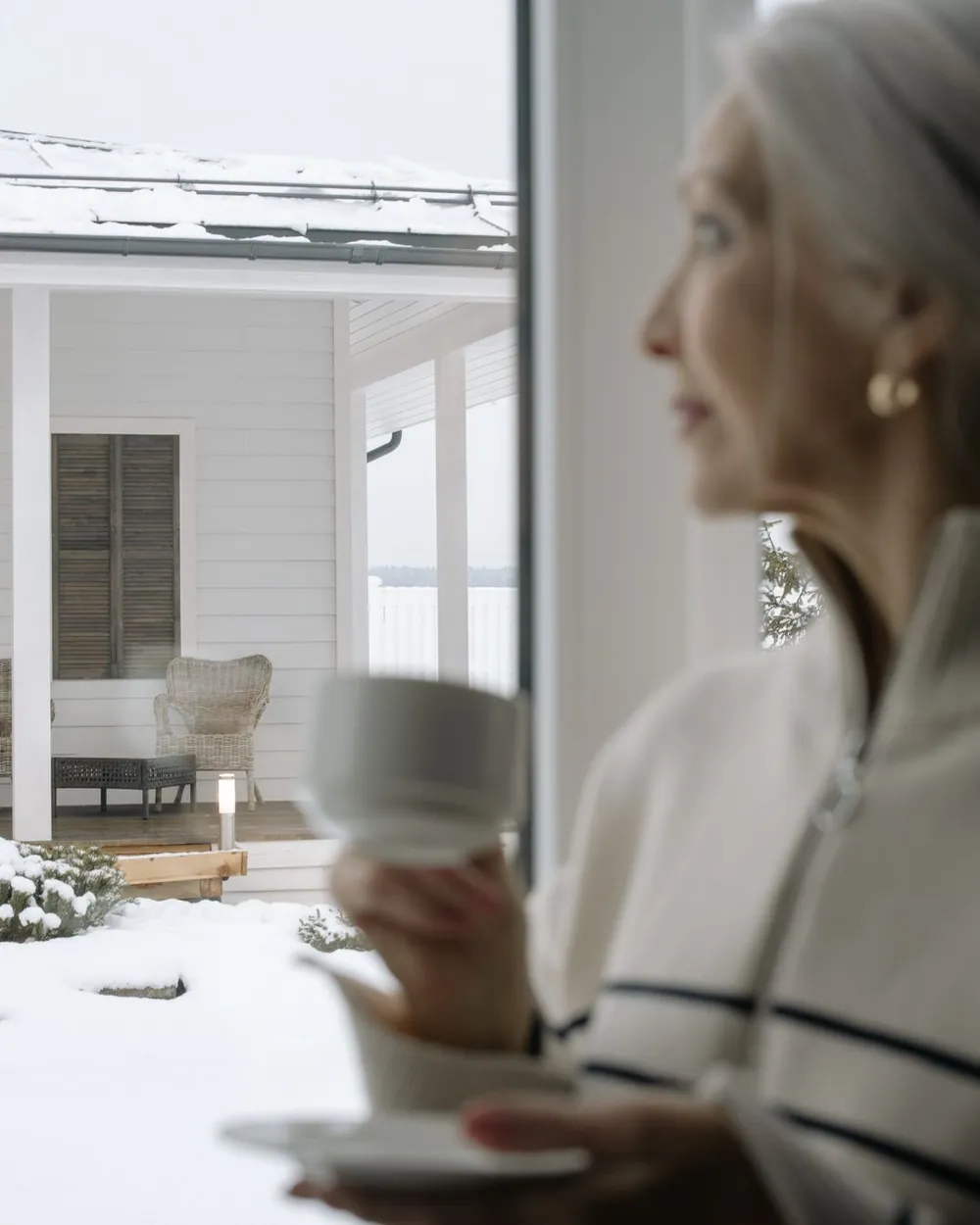 Oudere vrouw met een kop thee kijkt rustig door het raam naar een besneeuwde veranda