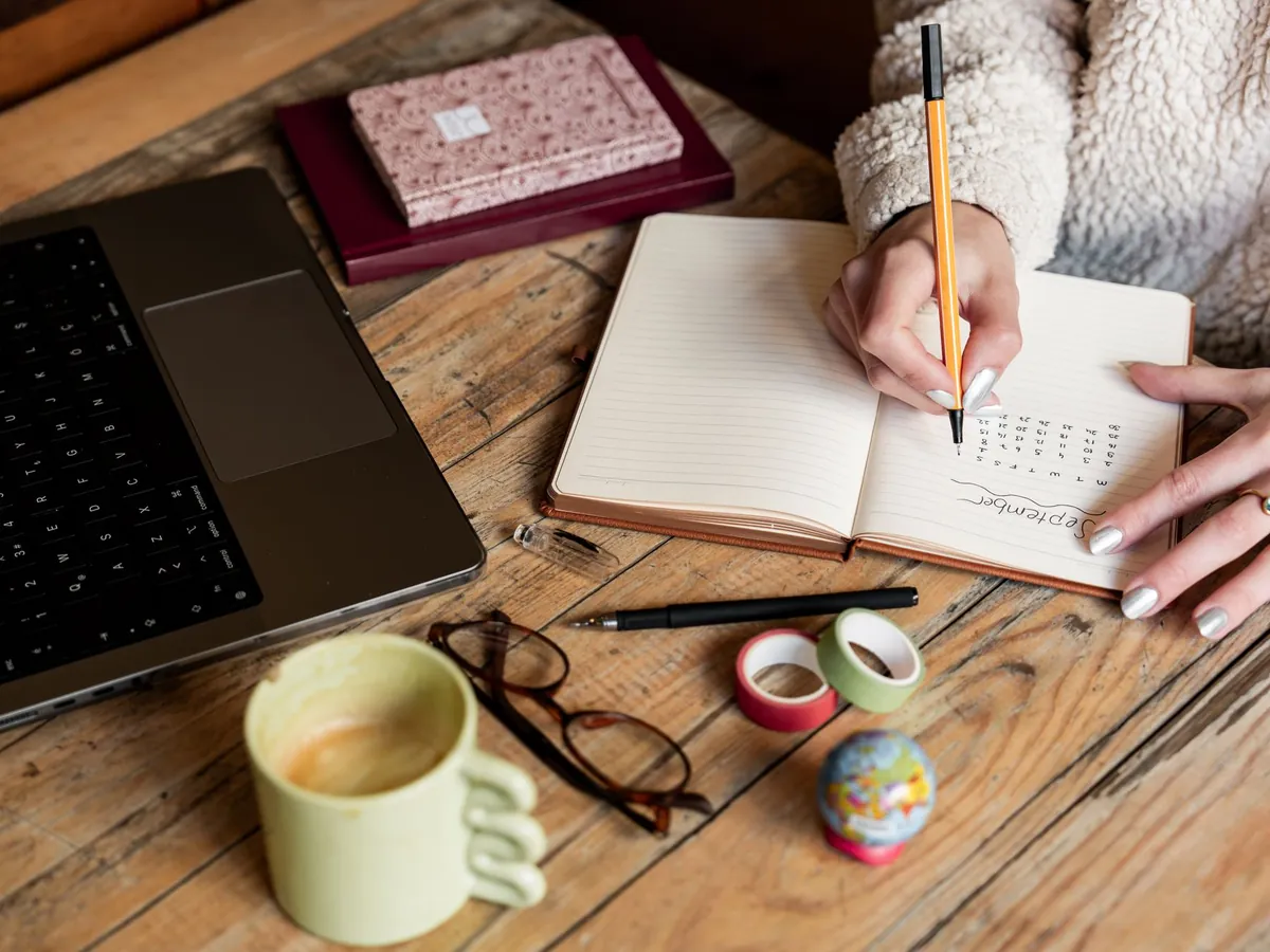 Vrouw schrijft in een planner naast haar laptop en een kop thee op een houten tafel