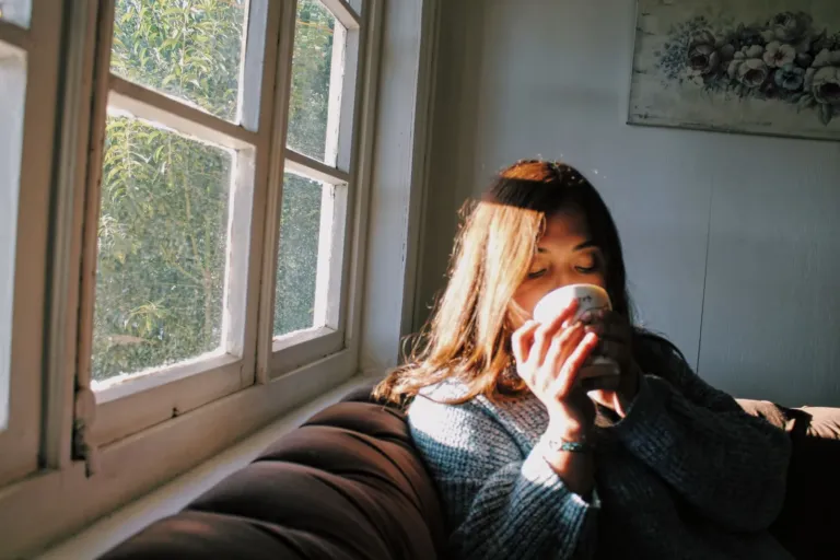 Vrouw zit op de bank bij het raam en drinkt uit een kopje in zacht daglicht