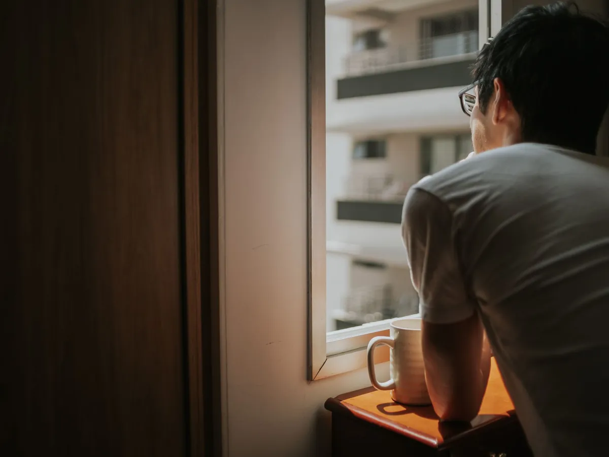 Man met bril staat bij het raam met een mok en kijkt naar buiten