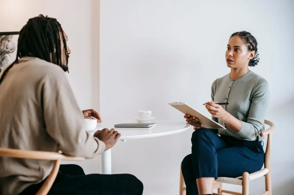 Begeleidster met klembord in gesprek met een man aan een kleine ronde tafel