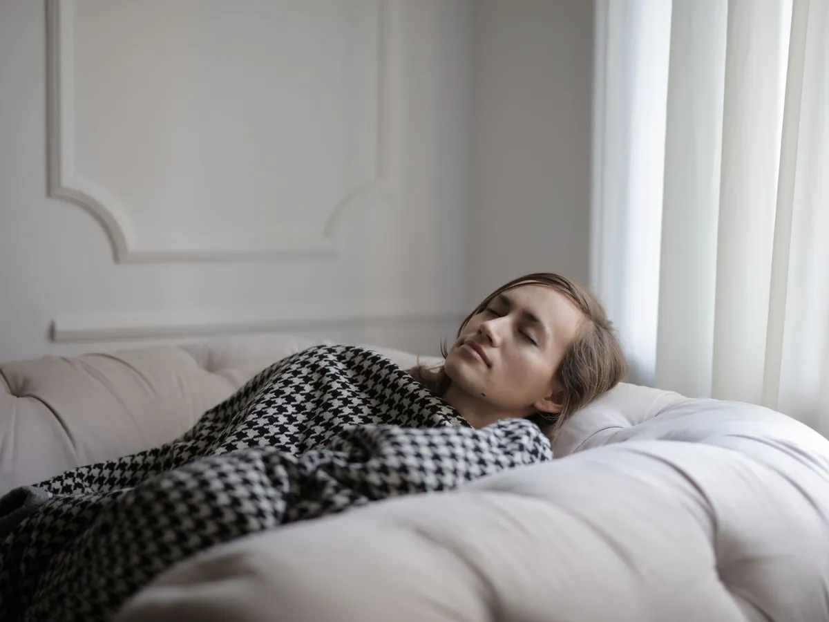 Vrouw rust met gesloten ogen op een lichte bank onder een geruite deken